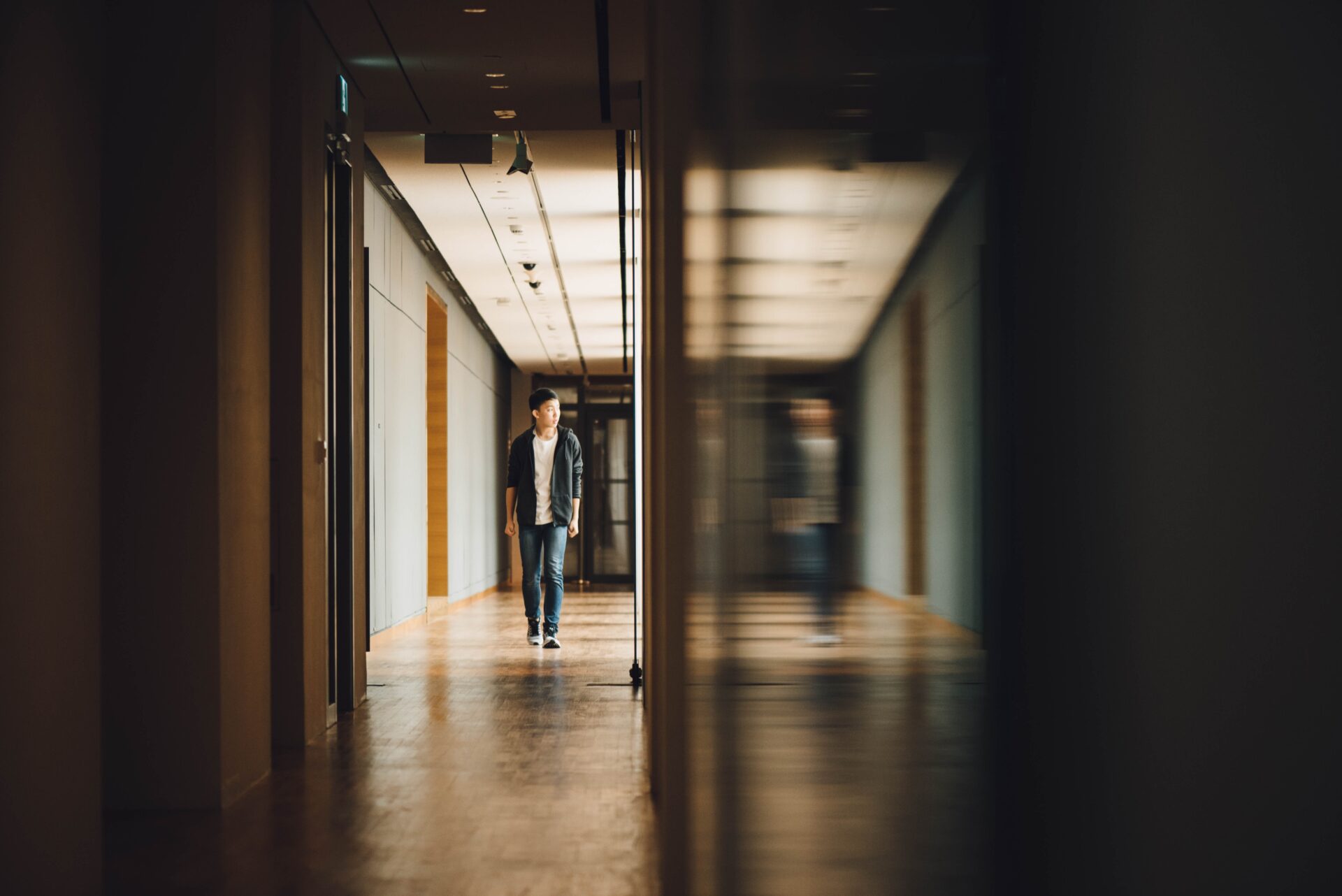 a boy walking alone down a hallway towards the camera, his blurry reflection on the right