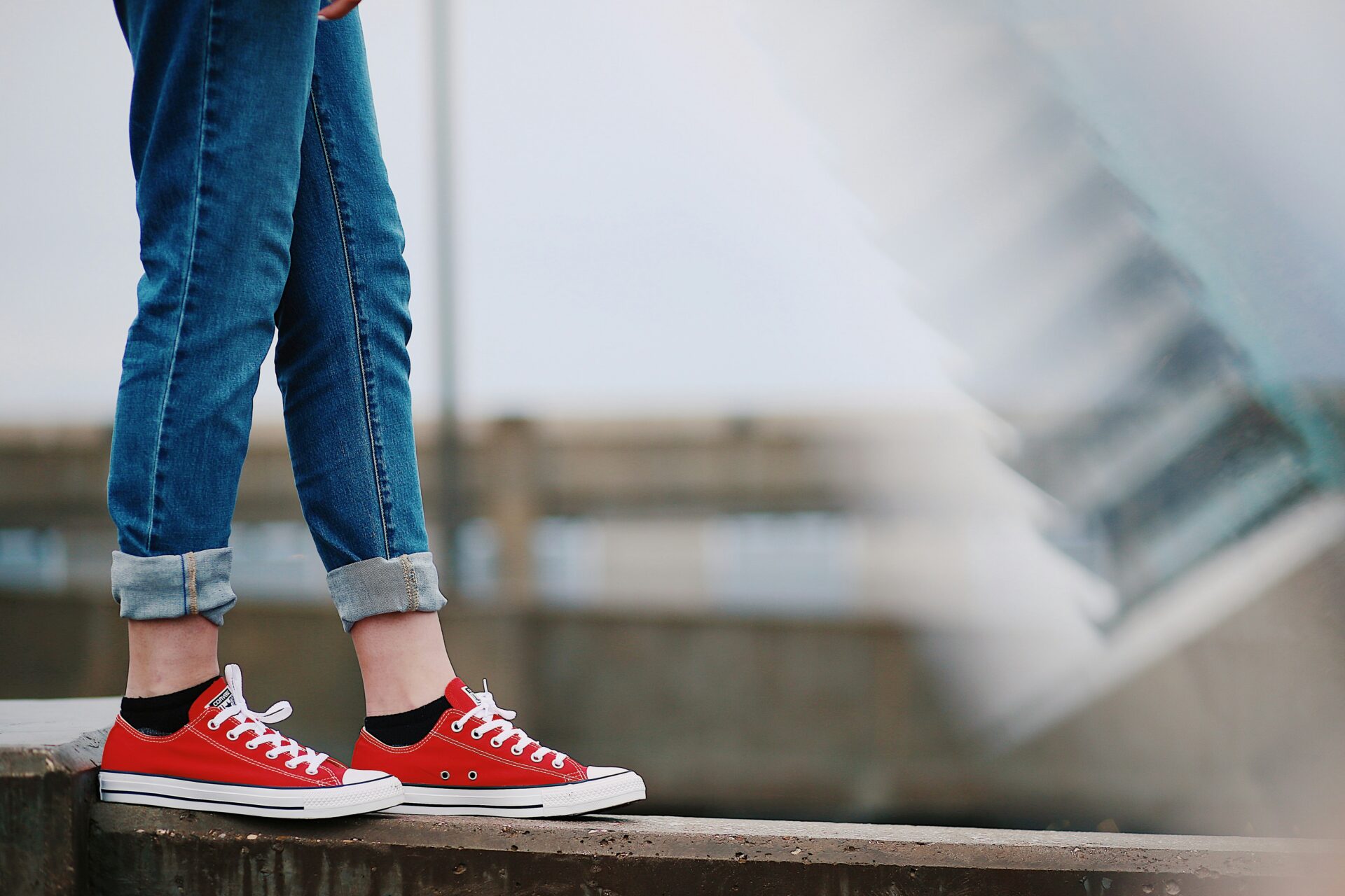 A young person in jeans with a rolled hem and red converse sneakers walks along the edge of a concrete ledge.